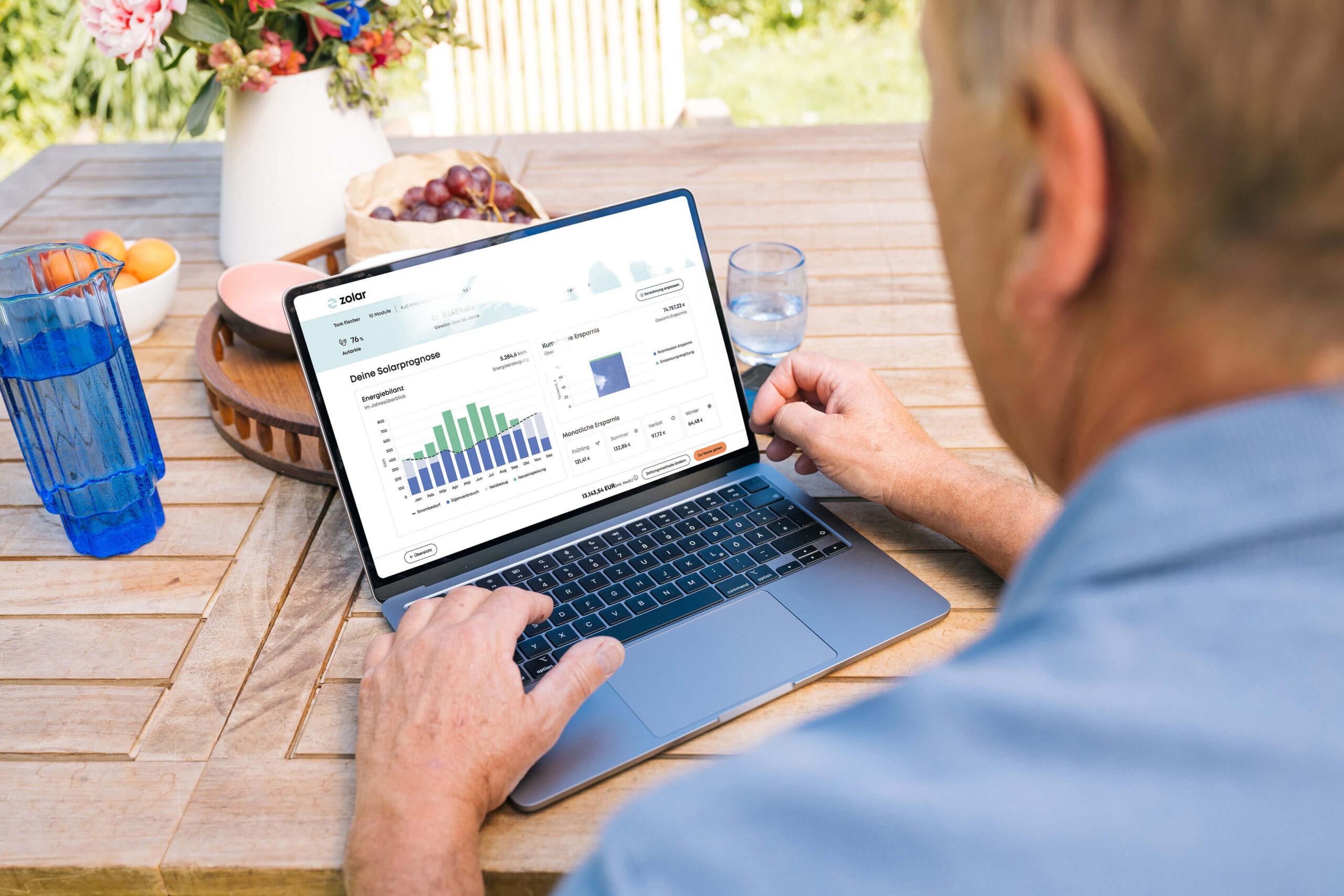 Blick über die Schulter eines Mannes, mit Blick auf einen Laptop auf dessen Bildschirm man den zolar Online Konfigurator sieht und ein Dashboard mit Daten zur Solarprognose für die Konfiguration. Der Laptop steht auf einem Gartentisch, neben frischem Obst, einem Blumenstraß und Wasser.